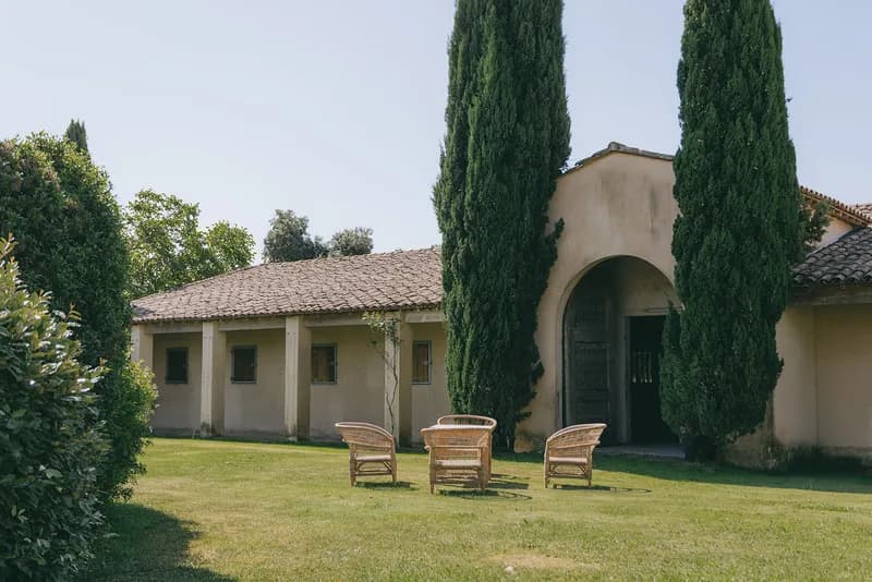 Rattan armchairs on the lawn before a building with an arched porch and cypresses